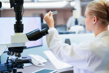 Caucasian female scientist inspecting glass slide near microscope in modern lab. Young researcher...