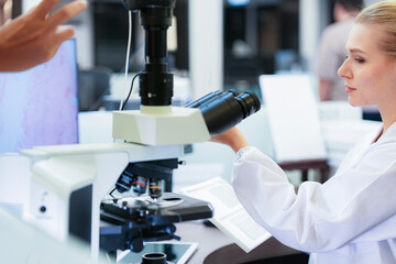 Serious Caucasian female scientist inspecting glass slide near microscope in modern lab. Young researcher checking histology sample with digital pathology monitor background.
