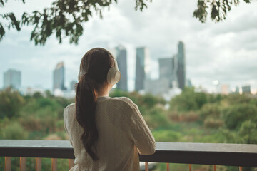 Rear view of young asian woman wearing headphones looking at blurred city skyline and green nature for relaxation and modern urban lifestyle