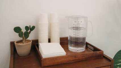 Water refill station with reusable cups, measuring jug and napkins on wooden tray. Concept of self service hydration, sustainable lifestyle, cafe corner and minimal interior design.