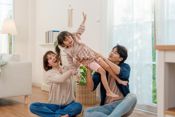 Joyful Asian parents sitting on floor, lifting daughter high in bright living room. Family sharing playful, heartwarming moment of love and fun.