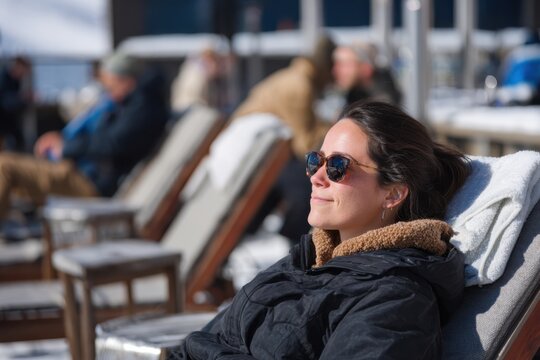 A woman in a black jacket and sunglasses reclines on a lounge chair at a sunny ski resort terrace, surrounded by people enjoying the bright winter weather in a relaxed setting