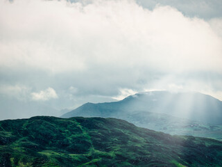 A mountainous landscape with sun rays streaming through clouds is shown. Green hills are visible in the foreground during the day. Taken near Coomloughra Horseshoe.