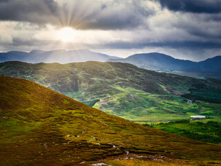 A scenic view of rolling hills and mountains against a dramatic sky. A bright ray of sunshine breaks through the dark clouds and illuminates the landscape of County Kerry.