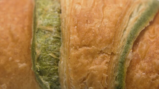 A macro video of a flaky croissant with visible matcha layers slowly rotating on a white background. The rotation highlights crisp texture, laminated dough, and green tea filling.