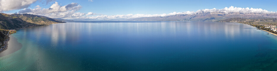 Ultra-wide aerial panorama of Lake Ohrid in Pogradec, Albania, showing calm blue water, distant mountains, and a dramatic cloud-filled sky.