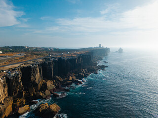 Aerial view of Cabo Carvoeiro coastal cliffs in Portugal, dramatic rocky shoreline above Atlantic Ocean with lighthouse, winding road and deep blue water