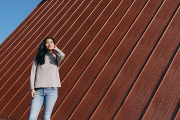 Woman standing beside a rusted metal wall in bright daylight, casual sweater and jeans, urban outdoor setting, confident stance, minimalist composition, texture and color contrast, street photography