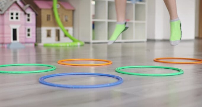 Small group of diverse children playing a hopscotch-style game with colorful plastic hoops in a sensory classroom, with a young boy with Down syndrome leading the activity while a teacher claps