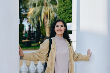 Woman stands between white columns outdoors, smiling, wearing a beige cardigan and light dress,...