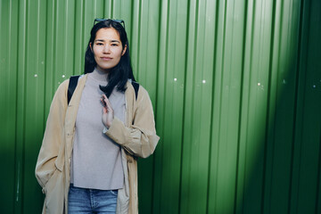 Woman in beige coat and gray scarf stands against a green metal wall, casual street style with...