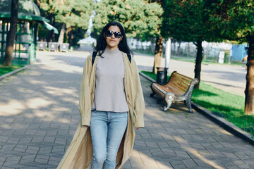 Fototapeta premium Woman walking in a park on a sunny day wearing a beige coat over a sweater, jeans, and sunglasses, showcasing casual outdoor fashion and street style against greenery and benches in a bright urban