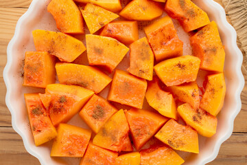 Pumpkin cut into pieces with sugar and cinnamon in a ceramic dish, ready to bake on a wooden table, close-up, top view.