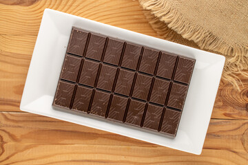 One bar of dark chocolate with a ceramic plate on a wooden table, close-up, top view.