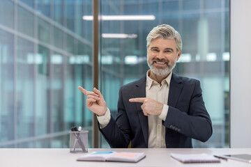 Experienced businessman sitting at an office desk, smiling confidently and gesturing to the left...