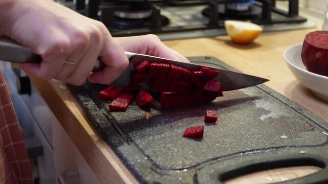 Close up of a person's hands slicing fresh raw beets with a sharp chef's knife on a dark cutting board in slow motion, highlighting healthy food preparation and home cooking in a cozy kitchen