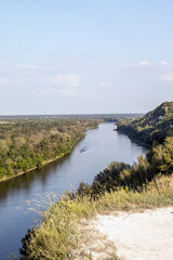 A river along a white chalk cliff and a green plain under a bright sky. A natural landscape with a wide river flowing along a high chalk cliff. To the left of the river is a green, wooded plain