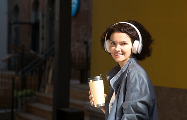 smiling middle aged woman with headphoneson drinks coffee the street of the city against the background of a yellow wall. International Music Day. Mindfulness and feel-good moments. Audio healing. 