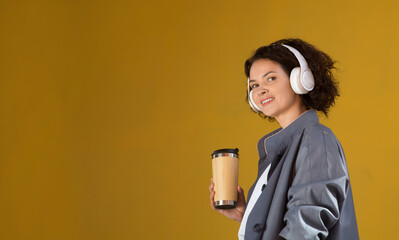 smiling middle aged woman with headphoneson drinks coffee the street of the city against the background of a yellow wall. International Music Day. Mindfulness and feel-good moments. Audio healing. 