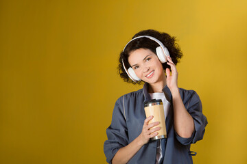 smiling middle aged woman with headphoneson drinks coffee the street of the city against the background of a yellow wall. International Music Day. Mindfulness and feel-good moments. Audio healing. 