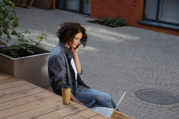  remote workers. Working from Anywhere.  middle aged woman    is working on a laptopon and talking on a mobile phone a bench on the street in a cafe. startup