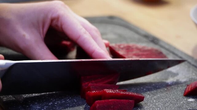Close up of a person's hands slicing fresh raw beets with a sharp chef's knife on a dark cutting board in slow motion, highlighting healthy food preparation and home cooking in a cozy kitchen