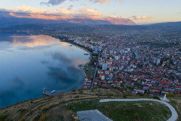 Aerial view of Pogradec, Albania, at sunset, showing the city skyline along Lake Ohrid with calm water and mountain reflections.