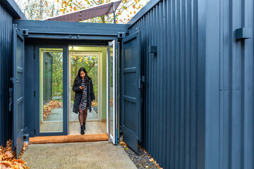 Businesswoman leaving of modern shipping container coworking, checking her cellphone, an innovative workspace powered by solar energy. Contemporary sustainable and eco friendly architecture concept.