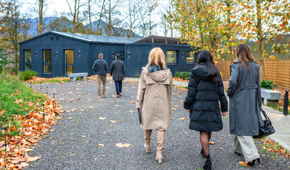 Unrecognizable real estate agent walking with investor group to modern passive house built from shipping containers in a corporate visit to sustainable construction, surrounded by autumn leaves