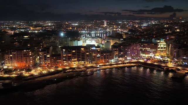 Cinematic night aerial strafes along the vibrant Sliema and St. Julian's coastline, revealing the glowing urban skyline and city lights reflecting on the Mediterranean Sea in Malta