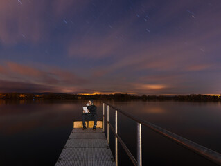 Obraz premium Man is using, working on tablet device sitting on floodgate at starry night. He is working remotely