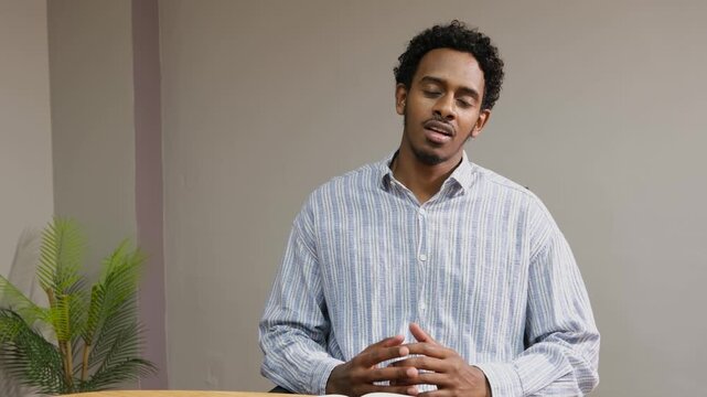 Young black man wearing a striped shirt speaking confidently during an online video conference or remote business meeting from a modern office setup.