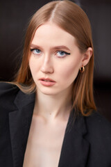 A woman in a black jacket and black trousers is sitting on a brown sofa. She has long brown hair and trendy makeup.