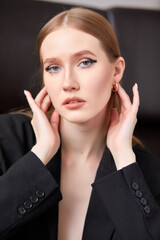 A woman in a black jacket and black trousers is sitting on a brown sofa. She has long brown hair and trendy makeup.