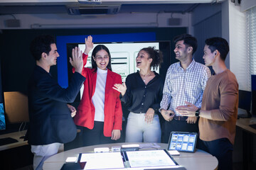 Diverse group of professionals celebrating successful teamwork high-fiving and smiling enthusiastically around a table with tablets displaying app designs. Collaboration, innovation, and digital strat