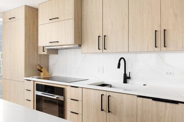 A kitchen detail with a black faucet on a white marble countertop, stovetop and stainless steel oven, and modern wood cabinets.