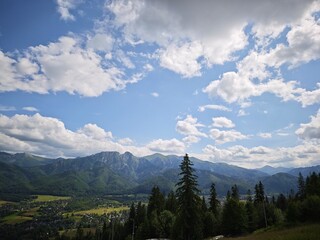 Massive mountains under cloudy blue sky above lush forest and distant town