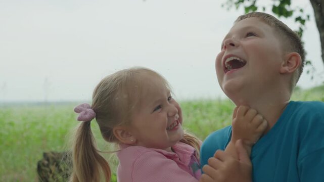 caucasian brother and sister laughing outdoors, big brother teases little sister under shady tree, both smiling and embracing, warm summer field backdrop, candid lifestyle shot capturing genuine joy