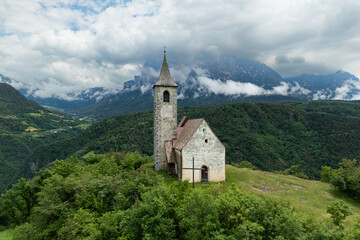 Fototapeta premium Chiesa di Sant Andrea in Renon above a hillside pasture with tree clusters, Catinaccio backdrop, aerial view. Dolomites region