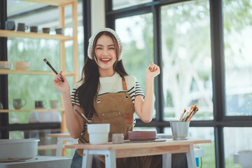 Happy young asian woman artist smiling while holding paint brush and working on pottery or ceramic artwork in cozy studio workshop