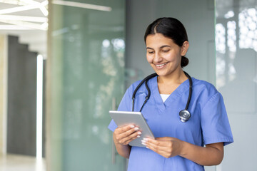Young happy female doctor or nurse in medical scrubs and stethoscope smiling while using a digital tablet, for patient care and data management in a modern hospital setting