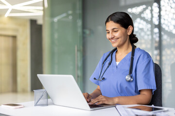 Young female medical professional wearing scrubs and a stethoscope, smiling while typing on a laptop at a desk, researching patient data for remote consultation in a modern hospital setting