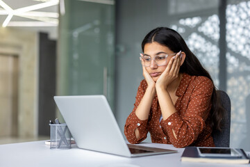 Young indian woman feeling tired, bored, and overwhelmed, resting her face in her hands while looking at a laptop screen in a modern office, experiencing work burnout