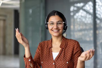 Young woman wearing a headset and spectacles, smiling friendly while working in an office,...