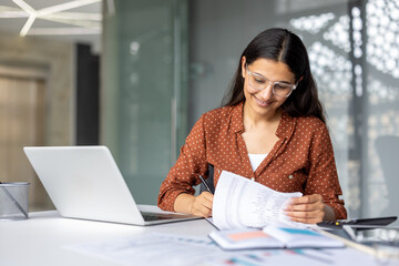 Indian woman in glasses and spotted shirt at office desk, smiling while reviewing documents, writing notes and working on a laptop in a modern, focused workplace