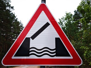 Road Narrows Bridge Warning Traffic Sign Against Green Forest. Triangular red road sign warning of narrowing bridge ahead, set against lush green forest background. Road safety and traffic concept.