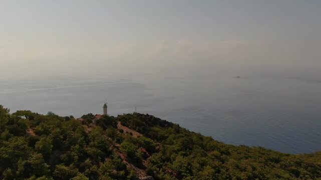 A serene, wide drone shot of a traditional lighthouse standing atop a hill covered in greenery. An endless, misty sea view stretching into the horizon. Gelidonya Lighthouse.