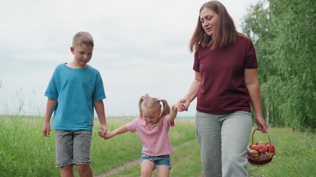 white family walking meadow path holding hands mother carries basket of apples while smiling brother and little sister step along grassy country lane under soft overcast sky warm bonding, playful