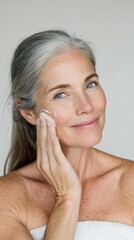 Mature woman with gray hair smiling, applying moisturizing cream to her cheek for skin care, focusing on beauty ritual, well being, and anti aging routine