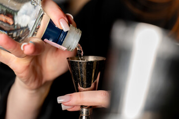A person is pouring a clear liquid from a small glass bottle into a metal jigger at a bar, preparing a cocktail or mixed drink.